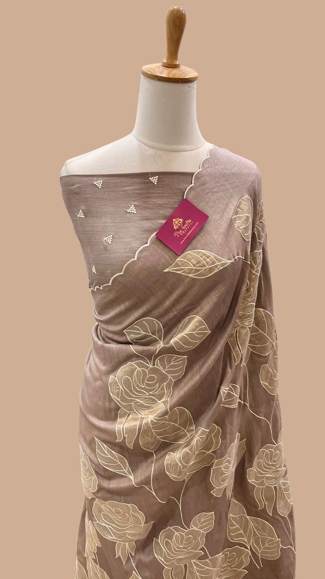 A draped taupe-brown saree with beige floral embroidery, displayed on a white mannequin torso against a solid beige background.